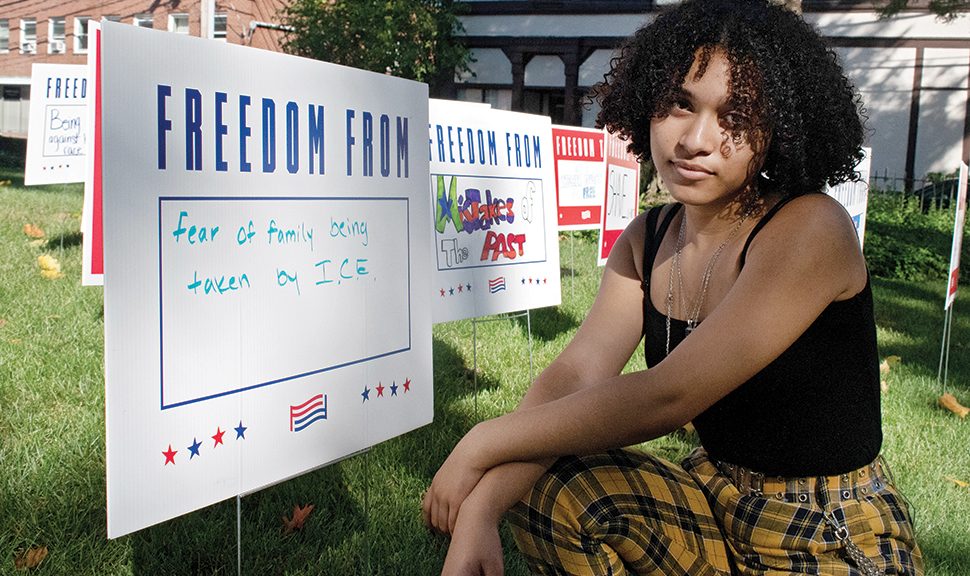 Teen leader Iris Alvarenga poses in front of yard signs at Waltham (Mass.) Public Library that depict issues youth patrons care about. The installation was a partnership between the library, civic organization For Freedoms, and local art group Blueprint Projects. Photo: Erwin Cardona/Waltham (Mass.) Public Library