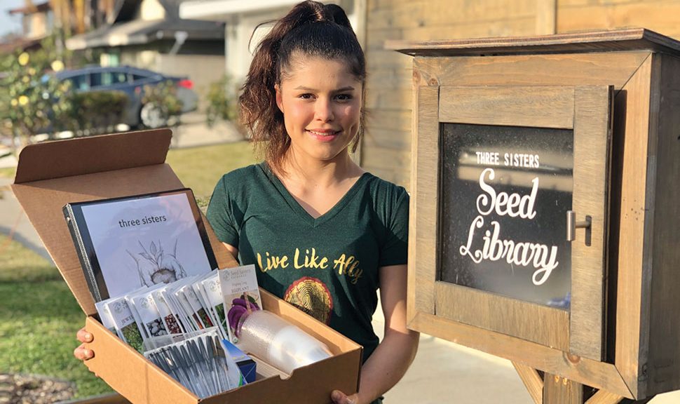 Alicia Serratos poses in front of a seed library holding a box of seeds.