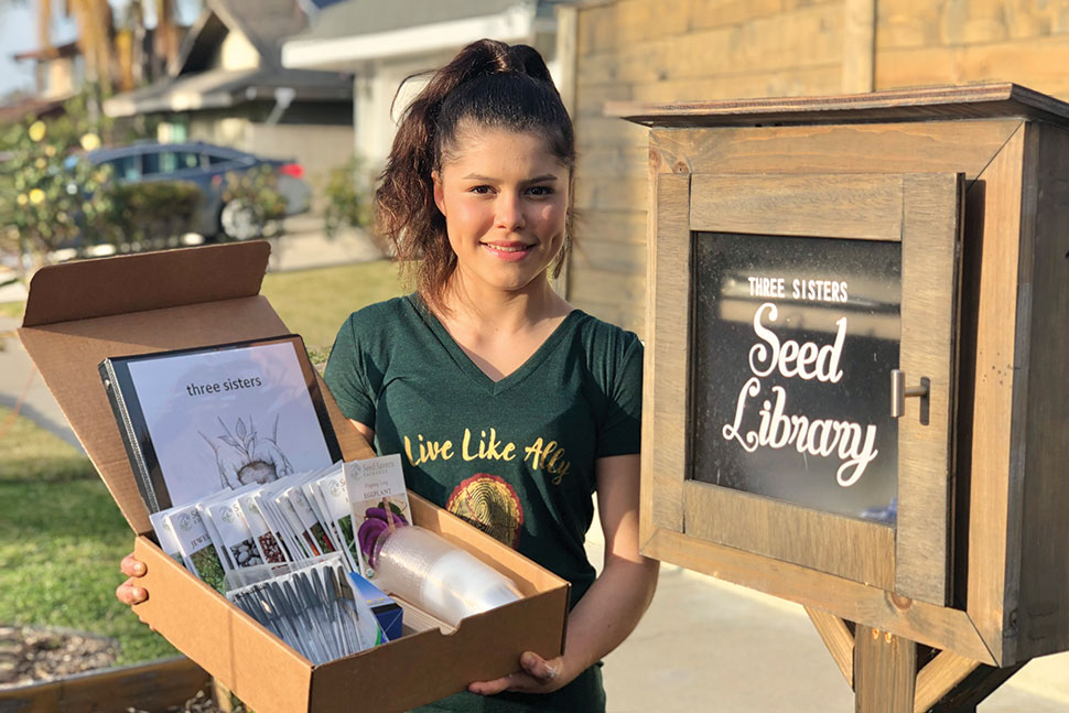 Alicia Serratos poses in front of a seed library holding a box of seeds.