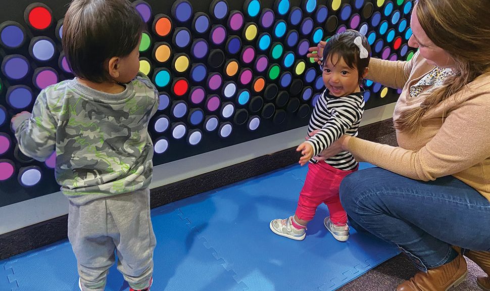 Two toddlers and an adult play with Everbright, an interactive light wall with many multicolored round dials. The smallest child faces the camera smiling.