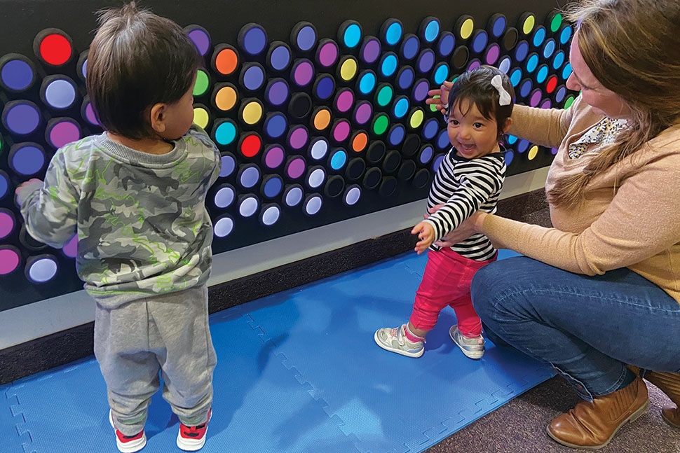 Two toddlers and an adult play with Everbright, an interactive light wall with many multicolored round dials. The smallest child faces the camera smiling.