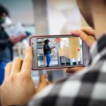 Man in plaid shirt, back to camera, films library patron with a cellphone