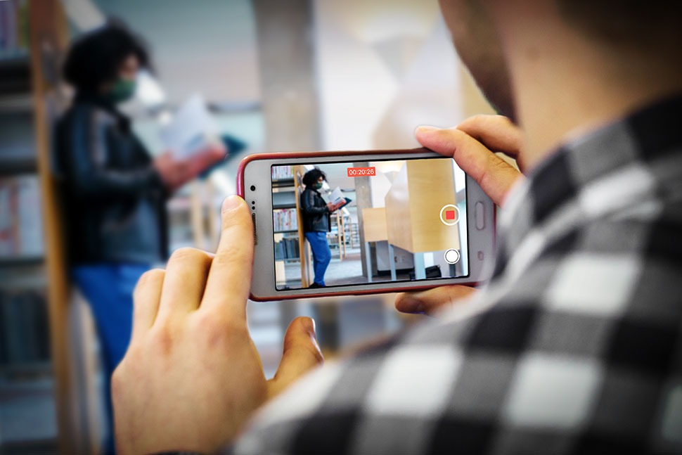 Man in plaid shirt, back to camera, films library patron with a cellphone