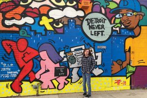 Photo of Cameron Socha, who graduates this year from Wayne State University’s School of Information Sciences, posing in front of a mural in Detroit. He collaborated with his professor Joan Beaudoin to catalog local murals.