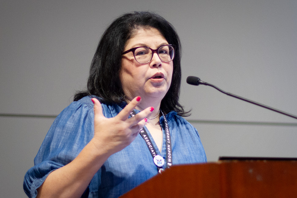 Becky Calzada wearing a blue blouse, glasses and holding right hand in air as she speaks into microphone