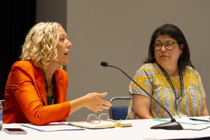 Pictured: Erin MacFarlane wearing an orange jacket and has yellow hair speaks into microphone as Becky Calzada, wearing glasses with dark hair and a yellow shirt, listens