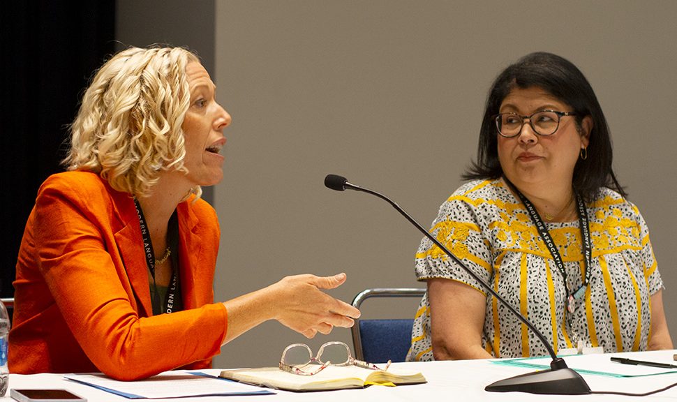 Pictured: Erin MacFarlane wearing an orange jacket and has yellow hair speaks into microphone as Becky Calzada, wearing glasses with dark hair and a yellow shirt, listens