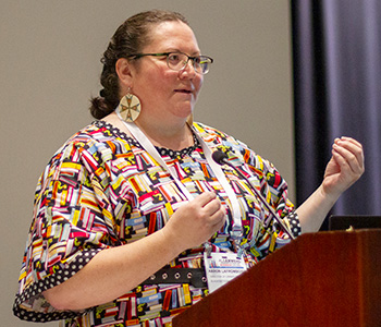 Aaron LaFromboise, director of library services at Blackfeet Community College in Browning, Montana, is shown speaking at a podium, wearing glasses, earrings, and a patterned dress.