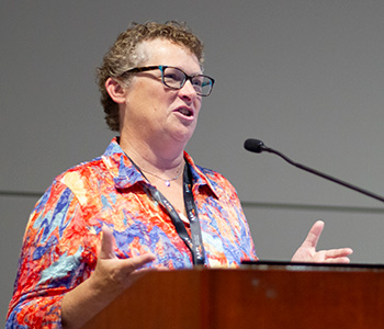 Val Edwards, library teacher leader at Madison (Wis.) Metropolitan School District, is wearing glasses and an orange and blue top while she speaks at a podium. 