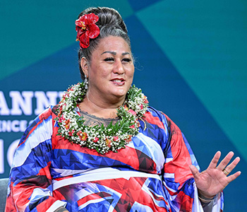 Native Hawaiian teacher, author, and filmmaker Hinaleimoana Wong-Kalu is seen on the speaker stage wearing a red flower in her hair, a necklace made of flowers, and a red, white, and blue dress.