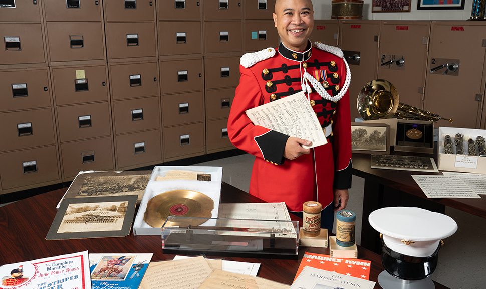 Philip Espe stands in a room with filing cabinets in the background. He is in a red US Marine Band uniform holding a stack of sheet music. Various pieces of music, memorability, and children's books are arranged on the table in front of him.