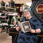 Anna Kresmer, holding a title from the B&O Railroad Museum research library, poses in front of a 19th century locomotive.