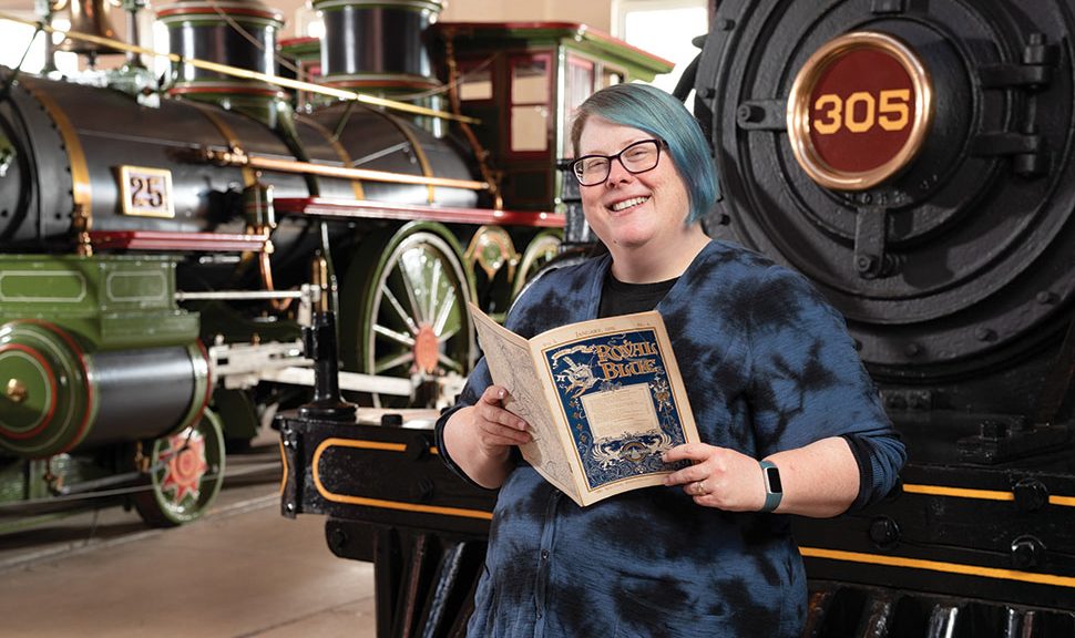Anna Kresmer, holding a title from the B&O Railroad Museum research library, poses in front of a 19th century locomotive.