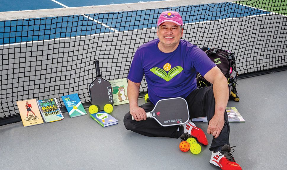 Drew Evans seated on a pickleball court with books about pickleball propped up on the net