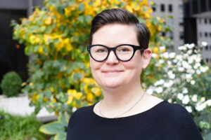 Headshot of Kerri Price, taken outside, with trees in the background. She is wearing black and has short hair and glasses.