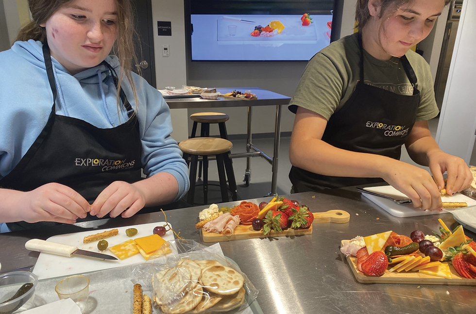 Two teens wearing aprons work side-by-side at a metal counter, arranging an array of cheese, meat, fruit, vegetables, and crackers on their boards.