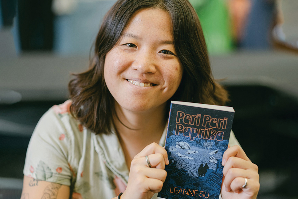 Author Leanne Su holds up a paperback book with a blue cover and the title and her name in yellow.