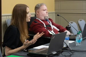A woman with long hair seated at a table next to a bearded man with a ventilator gestures as she speaks. Both have laptops open in front of them on the table.