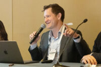 A smiling man wearing a suit, seated next to other panelists at a table, speaks into a microphone in his hand.