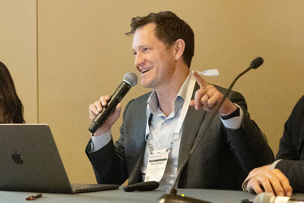 A smiling man wearing a suit, seated next to other panelists at a table, speaks into a microphone in his hand.