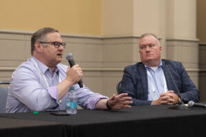 Seated at a table, a man in a patterned shirt and glasses speaks into the microphone he's holding and gestures with the other hand, while a man in a suit next to him watches him intently.