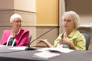 Two women with white hair—one wearing pink, one wearing yellow—sit together at a conference table with a microphone. One speaks, while the other listens and watches her.