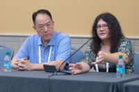 A woman seated at a table in front of a microphone speaks to the audience. A man, one of her co-presenters, looks at the ahead and listens with his hands folded in front of him.