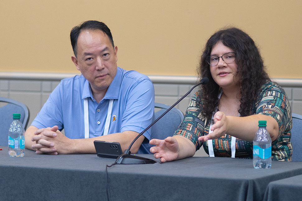 A woman seated at a table in front of a microphone speaks to the audience. A man, one of her co-presenters, looks at the ahead and listens with his hands folded in front of him.