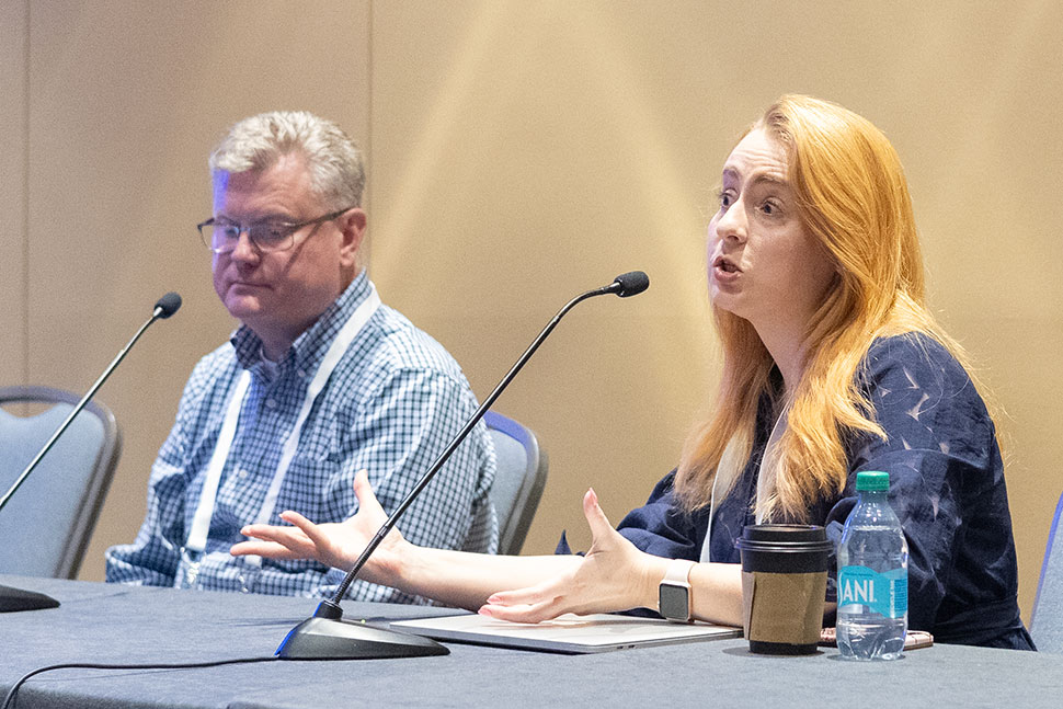 A man and a woman sit in front of tabletop microphones. The woman, Annette Bochenek, is speaking. Next to her are a takeout cup of coffee and a water bottle.