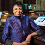 Carla Hayden in purple suit jacket, standing with elbow on a low library bookcase