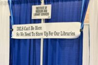 Empty conference booth with a sign that reads: IMLS Can't Be Here So We Need to Show Up For Our Library