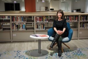 Librarian and archivist Colleen Barbus is wearing black and seated in a blue comfy chair in front of rows of low bookshelves making up the ALA Library